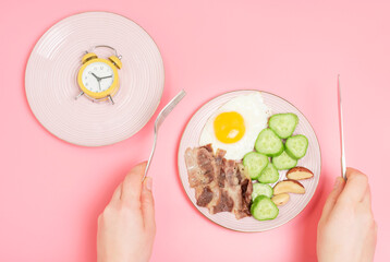 Hands holding fork and and knife next to plates with an alarm clock and food on a pink background.