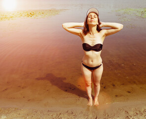 A young woman in a black swimsuit and white cap raises her head up with her eyes closed while standing on the water near the beach. Relax woman portrait. brown tint. The sun is shining