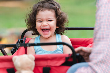 Fototapeta na wymiar Portrait of little Caucasian cute child girl smiling laughing and looking at small bear doll holding by father. adorable daughter sitting in trolley have fun playing with young dad at park on summer