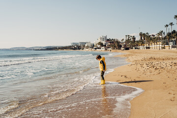 Boy in yellow rubber boots playing with water and sand at the beach. School kid touching water at...