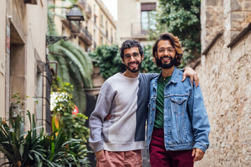 Naklejka premium gay male couple embracing looking at camera smiling happy in a nice street with plants, concept of leisure and love between people of the same sex