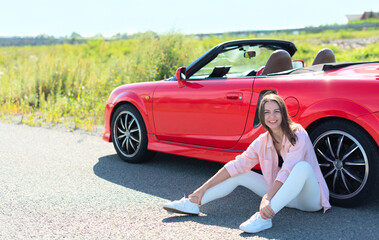 Lifestyle portrait of a carefree beautiful woman dressed casually sitting near red cabriolet car on the roadside. Road trip enjoying freedom concept 