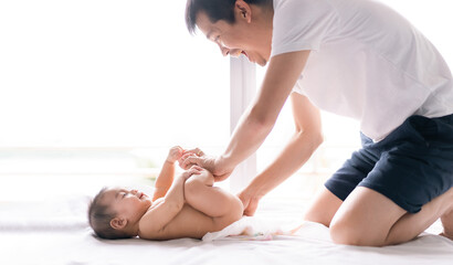 Asian father is changing diapper for his little baby boy in the morning time and the boy express happy moment with his smiley face, concept of role of father to care their child in family life. - obrazy, fototapety, plakaty