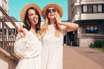 Two young beautiful smiling hipster female in trendy summer white dress clothes and straw hats. Sexy carefree women walking in street. Positive models having fun, hugging and laughing