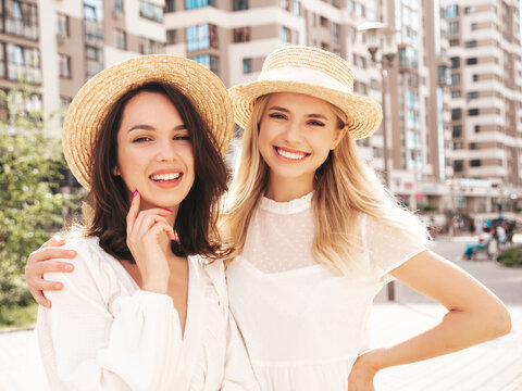 Two young beautiful smiling hipster female in trendy summer white dress clothes and straw hats. Sexy carefree women walking in street. Positive models having fun, hugging and laughing