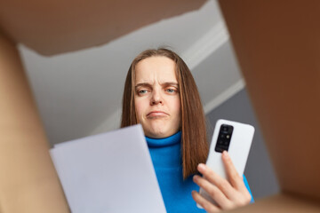Sad serious woman customer wearing blue turtleneck opening parcel, looking at consignment, trying...
