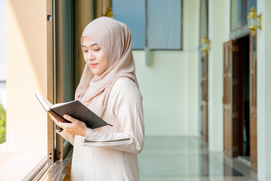 The image of an Asian Muslim woman in the Islamic religion in hijab in cream color. Standing reading the Qur'an and having a happy smiling face Staying in a beautiful mosque out of respect for God.