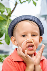 Toddler boy with panama grimaces with rubber bracelet on face near exotic plant closeup. Playful little child has fun looking in camera