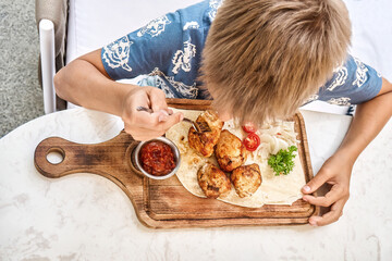 Blond boy eats grilled meat dish sitting at table in restaurant close upper view. Preteen child enjoys nutritious meal in cafeteria