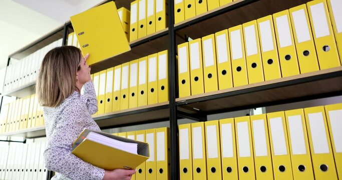 Woman accountant manager puts yellow folder on shelf with archive documentation