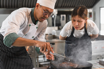 Portrait Asia young man in chef uniform cooking steak fire with student woman assistance chef at kitchen	