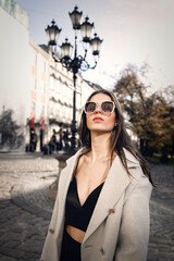 Fototapeta premium charming brunette posing on the street in spring, wearing a stylish beige coat, a top with razors and sunglasses