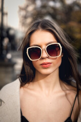 Fototapeta premium close-up portrait of a charming brunette on the street in spring, wearing a stylish beige coat, a top with razors and sunglasses