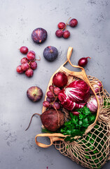 Food background with purple vegetables, roots and fruits in string shopping bag: radicchio, beetroot, red onion, grapes, figs. Harvesting, local farm market shopping, healthy food concept. Top view