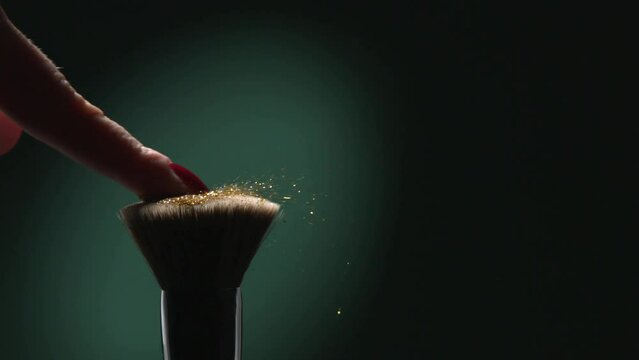 Woman Finger With Red Nail Flicks A Brush With Golden Glitter In Slow Motion On Green Aqua Menthe Color Background, Radiating Elegance, Beauty, And Creativity In Glamorous Makeup
