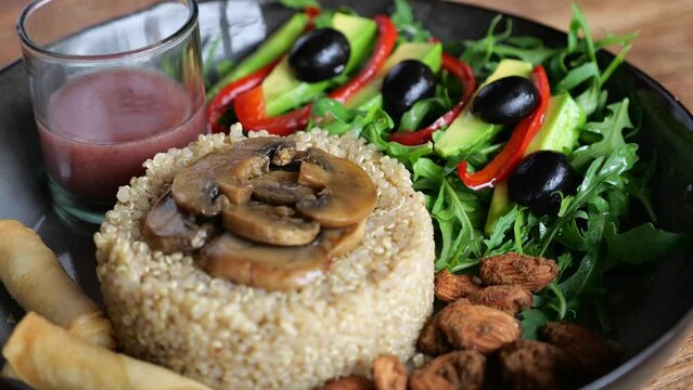 Dish on black plate: boiled rice with fried mushrooms on top, salad of avocado, bell pepper, olives, cilantro. Roasted almonds. Berry smoothie in small glass. Close-up of delicious nutritious meal.
