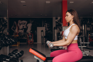 Fototapeta na wymiar Smiling sport woman training biceps with dumbbell in hand while sitting on bench in gym