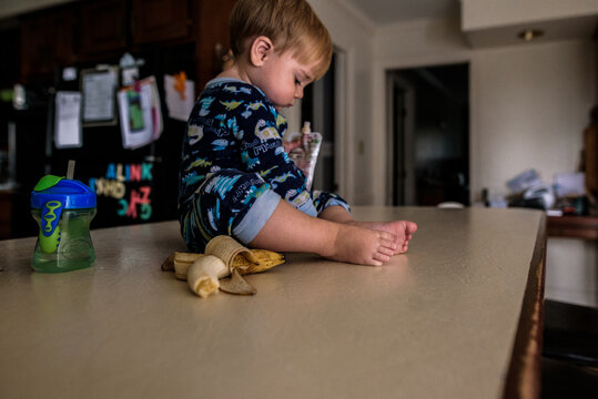 young boy sitting on kitchen counter with banana, cup, and pouch