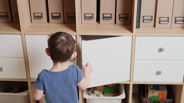 Toddler baby opens the closet door in the home living room. A small child opens a shelf drawer. Kid aged one year eight months