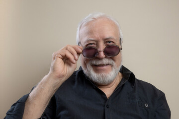 Obraz premium Charismatic elderly gray-haired man in sunglasses and gray shirt posing for camera in studio, looking with smile, close-up, light background