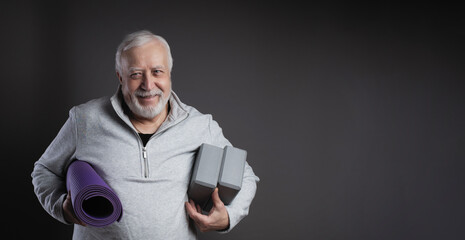 Charismatic elderly gray-haired man in tracksuit, mat and yoga support blocks, smiling in studio, gray background, space for text, banner
