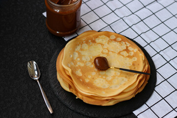Aromatic crepes or blinis are lying on black shale dish near a jar with condensed milk, super...
