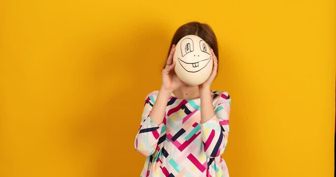Playful teenager girl covering her face of Ostrich egg with funny face on a bright yellow studio background, Face on the egg, celebrating Easter in style.