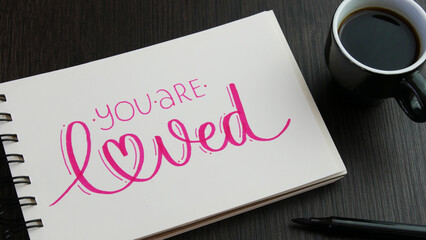 YOU ARE LOVED pink hand lettering in notebook with cup of espresso and pens on black wooden desk - obrazy, fototapety, plakaty