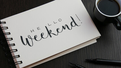HELLO WEEKEND! black lettering in notebook with cup of espresso and pens on black wooden desk