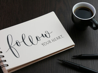 FOLLOW YOUR HEART. black lettering in notebook with cup of espresso and pens on black wooden desk - obrazy, fototapety, plakaty
