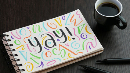YAY! black lettering with colorful motifs in notebook with cup of coffee and pens on black wooden desk - obrazy, fototapety, plakaty