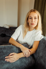 Serious blonde Caucasian girl sitting on sofa and looking curiously at camera