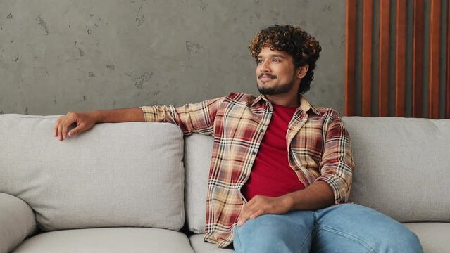 Young fun Indian man wears casual clothes look around think dream put hand prop up on chin lost in thought sits on grey sofa stay at home flat rest relax spend free spare time in living room indoor