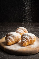View of three croissants on table with sugar falling, wooden table, selective approach, black background, vertical