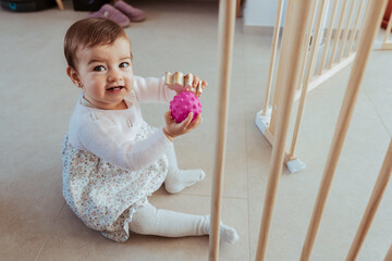 Child toddler playing with wooden and plastic toys at home, sitting on the floor, in front of child protection safety barrier fence. Cute baby girl smiling.