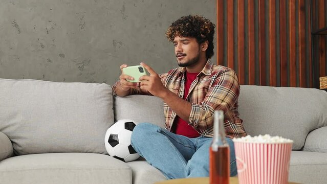Young sad fan Indian man wears casual clothes cheer up support football sport team use mobile cell phone sits on sofa stay at home flat rest relax spend free spare time in living room indoor grey wall