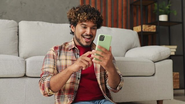 Young happy fun Indian man wears casual clothes hold use mobile cell phone do winner gesture sits on sofa couch stay at home hotel flat rest relax spend free spare time in living room indoor grey wall