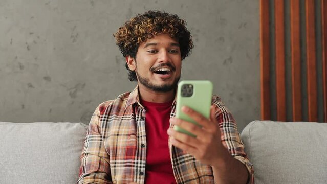 Young Indian man wear casual clothes get video call use mobile cell phone do selfie talk greet with hand sit on sofa couch stay home hotel flat rest relax spend free spare time in living room indoor