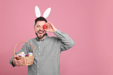 Happy man in bunny ears headband holding wicker basket with painted Easter eggs on pink background....