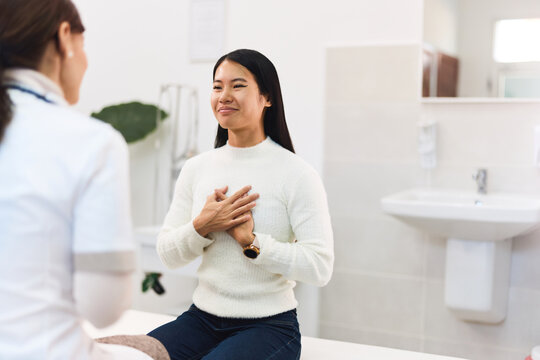 A Thankful Asian Patient Sitting On A Bed In The Examination Room With A Female Doctor.