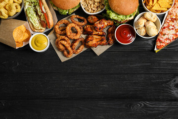 Burgers, onion rings and other fast food on black wooden table, flat lay with space for text