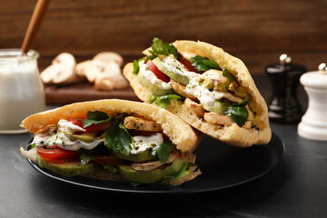 Delicious pita sandwiches with grilled vegetables and sour cream sauce on black textured table,...