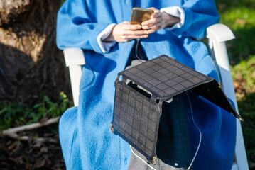 An unrecognizable woman with a mobile phone in her hands and a portable solar battery sits outdoors in the garden. Ecological alternative energy concept.