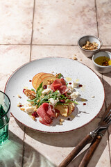 Salad of arugula and prosciutto or ham with the addition of sliced apple, cheese, walnuts on rustic plate in composition with cutlery and water glass on pink tile background. Copy space