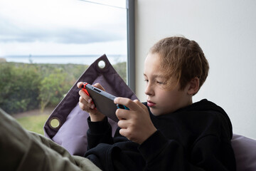 Leisure with games. Teenage boy relaxing on the balcony lying on beanbag bed and playing on handheld console. Child is focused on the action
