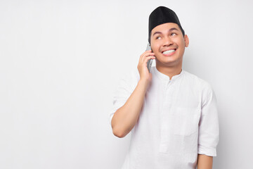 Portrait of smiling handsome young Asian Muslim man wearing Arabic clothes talking on mobile phone and looking empty space isolated on white background. People religious Islamic lifestyle concept