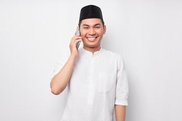 Portrait of smiling handsome young Asian Muslim man wearing Arabic clothes talking on mobile phone and looking at camera isolated on white background. People religious Islamic lifestyle concept