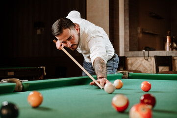 Handsome man is posing with billiard stick on the billiard table in the bar or salloon