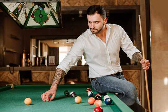 Handsome man is posing with billiard stick on the billiard table in the bar or salloon