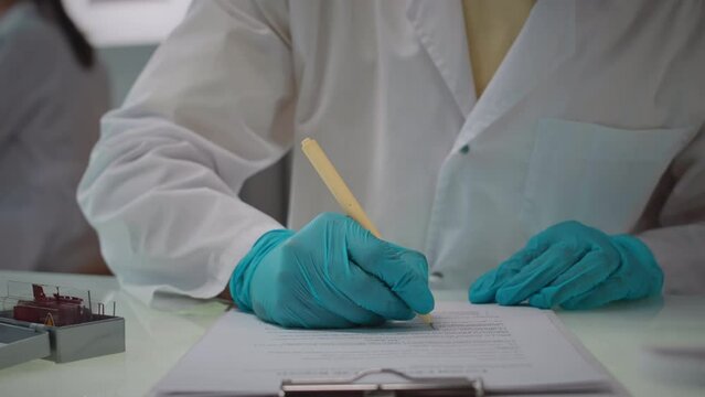 Cropped shot of unrecognizable scientist wearing white lab coat and gloves taking notes in scientific documents while doing research in laboratory